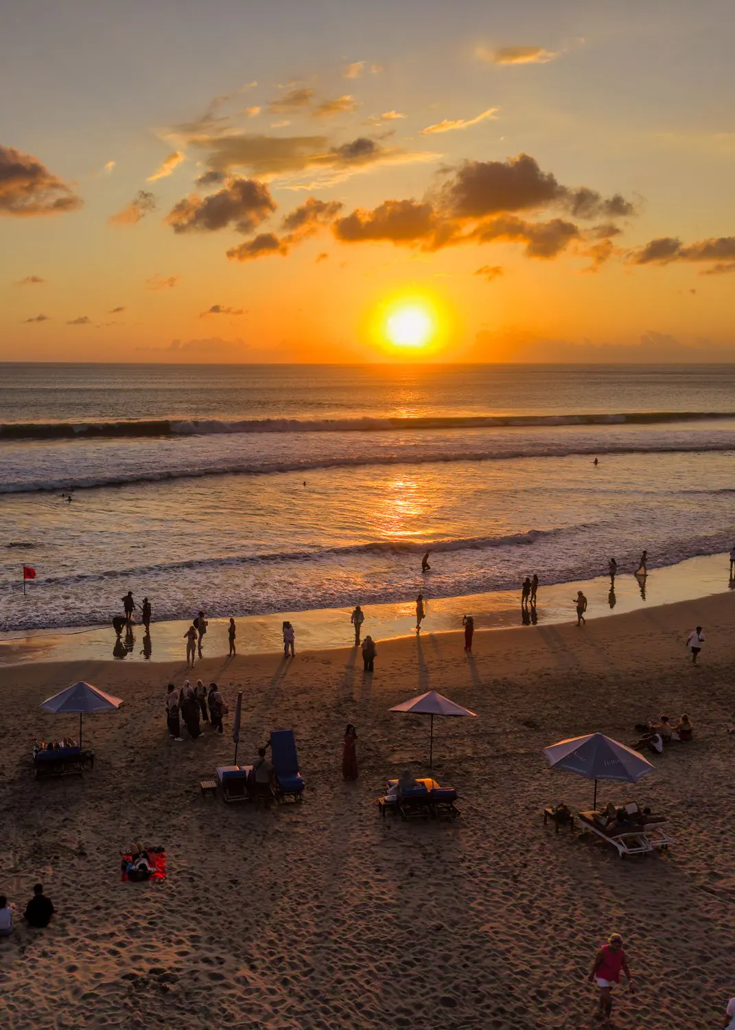 Sunset over the sea on Bali’s west coast, the sun low on the horizon and people on the sand among loungers and beach umbrellas