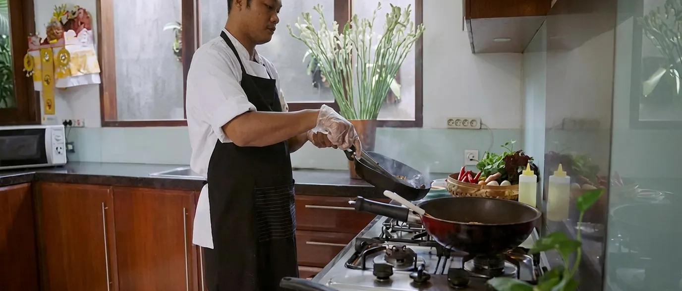 A chef at the gas range in the kitchen at Casa Brio, tipping a pan into a wok, with vegetables and a temple offering on the counter behind