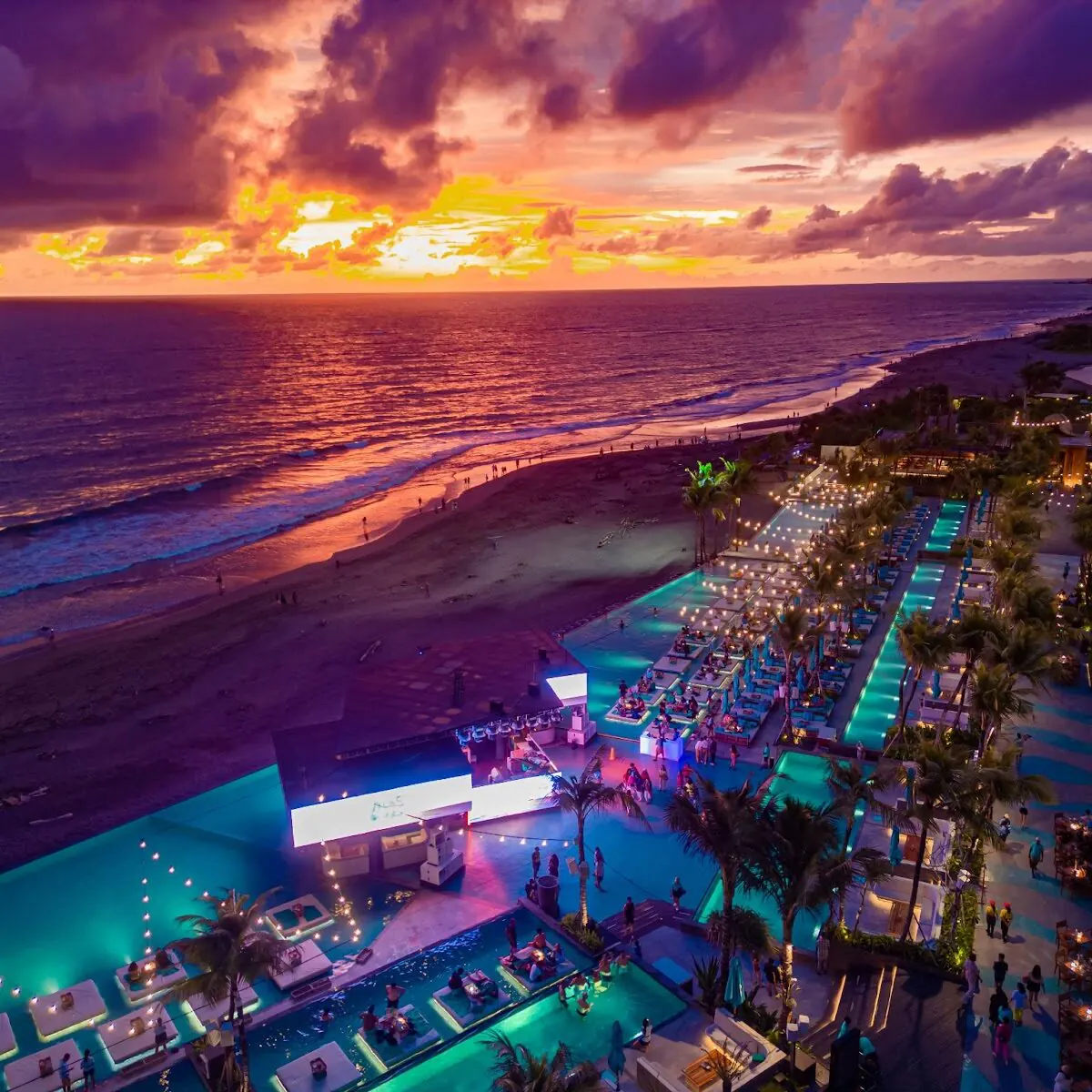 An aerial over a large Bali beach club at sunset, lit pools and rows of daybeds strung with festoon lights down to dark sand