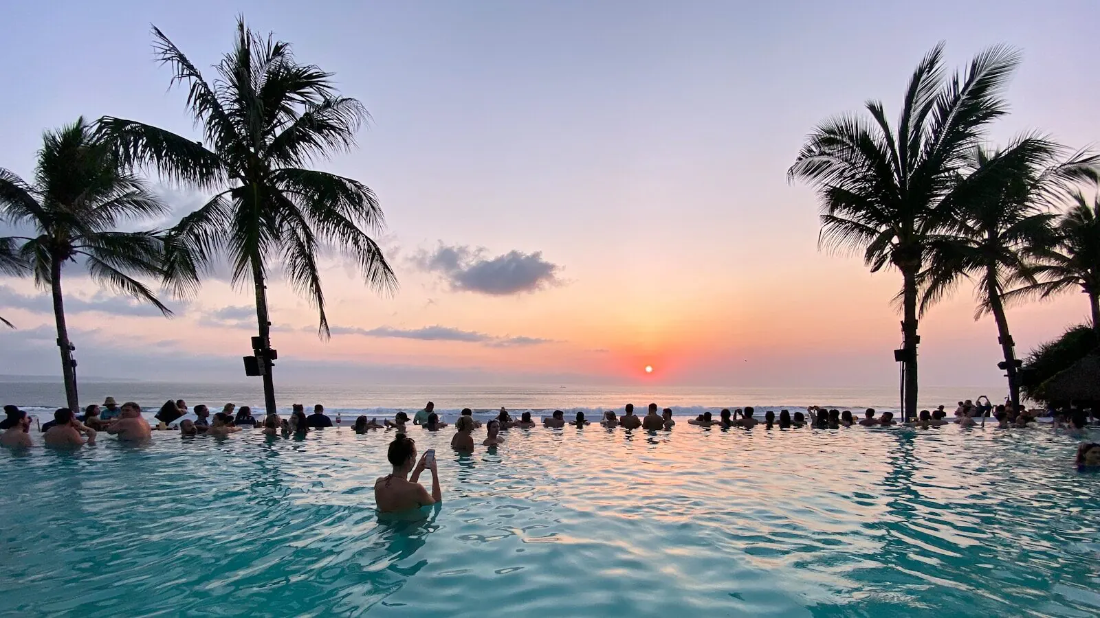 Sunset from inside the infinity pool of a Bali beach club, the far edge lined with people watching the sun drop into the sea