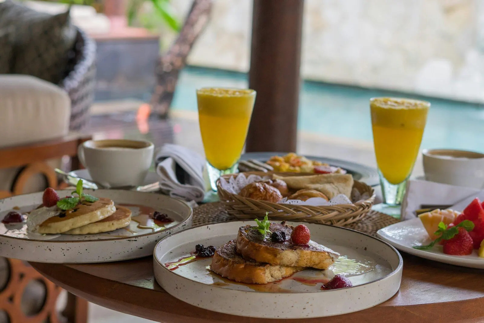 A poolside brunch table in Seminyak laid with cinnamon french toast and berries, pancakes, pastries, cut fruit and two glasses of juice