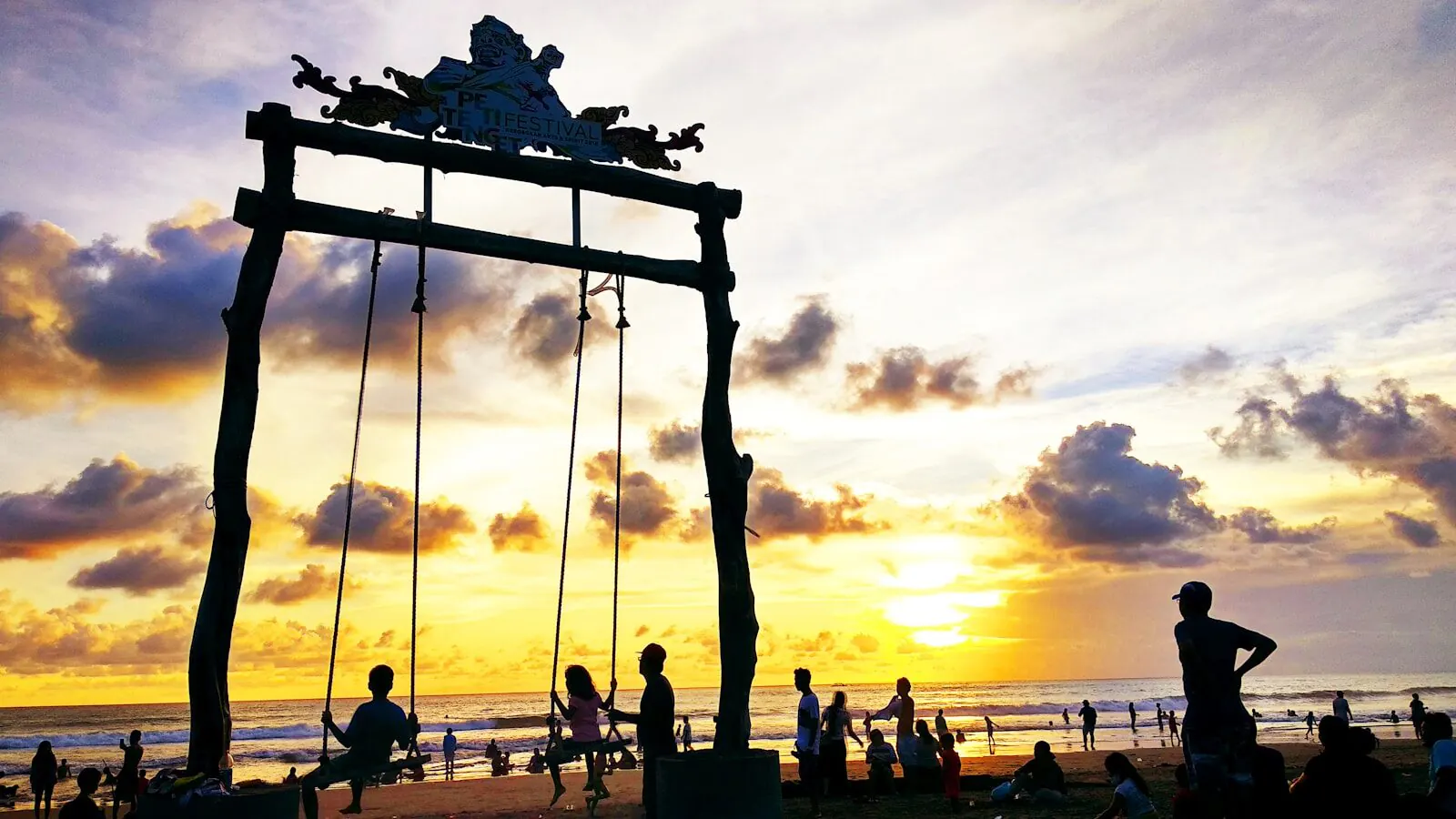 The tall timber swing on Petitenget Beach silhouetted against a gold sunset, its Petitenget Festival sign above the crowds in the shallows