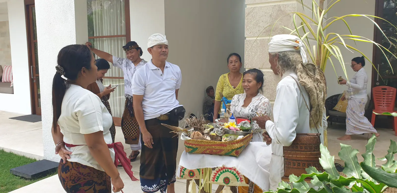 A pemangku in white ceremonial headcloth at the cloth-laid offerings table in the courtyard at Casa Brio, the house staff gathered round in kebaya