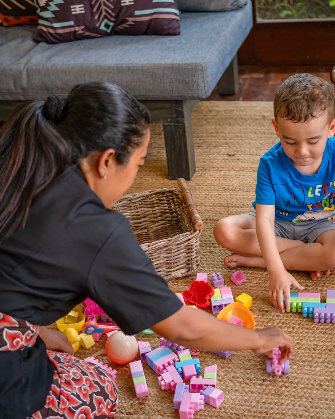 A member of the Casa Brio staff in the house uniform sitting on the jute rug beside a daybed, building coloured blocks with a young guest