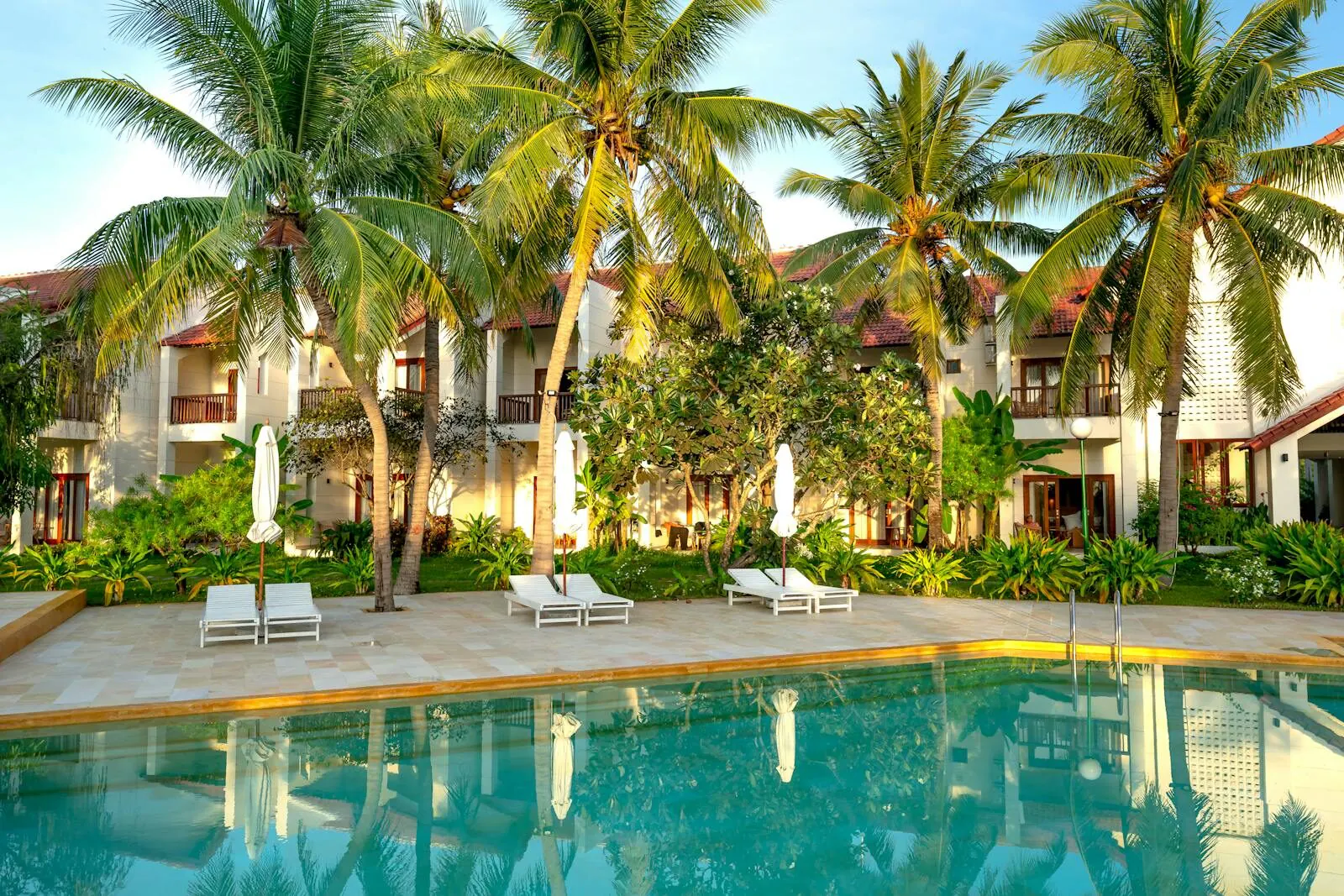A resort hotel pool at golden hour, white loungers and furled parasols on the terrace with two-storey guest blocks behind
